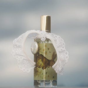 May include: A clear glass perfume bottle with a gold cap, featuring a label with a brown rabbit illustration and floral details. A white lace collar is tied around the neck of the bottle, accented by a single white oval charm hanging from the lace.