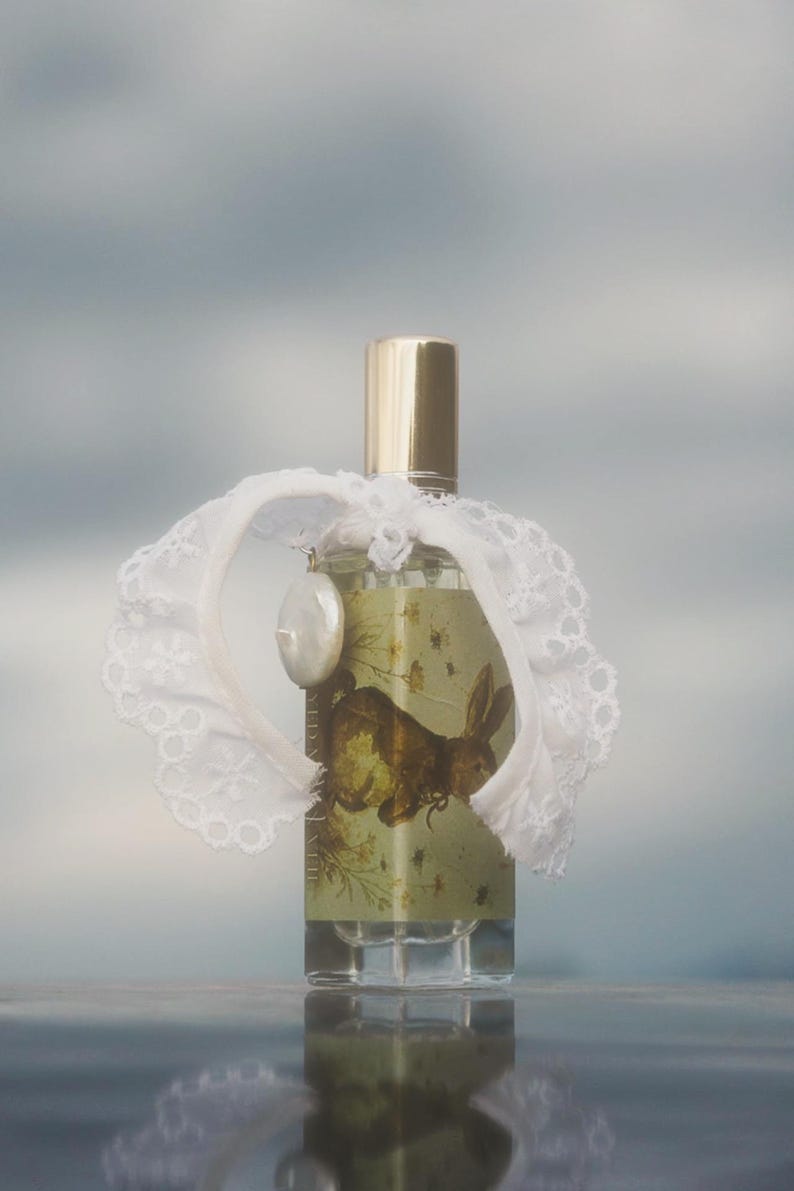 May include: A clear glass perfume bottle with a gold cap, featuring a label with a brown rabbit illustration and floral details. A white lace collar is tied around the neck of the bottle, accented by a single white oval charm hanging from the lace.