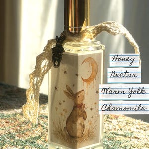 May include: A rectangular glass perfume bottle with a gold cap, featuring a label with a rabbit, crescent moon, and the text Honeyed Moon Veil. A cream-colored crochet ribbon is tied around the neck with a small metal charm. Text reads Honey Nectar, Warm Yolk, Chamomile.