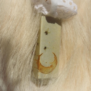 May include: A rectangular, light green glass bottle featuring a printed graphic of a golden crescent moon and several small flying insects. The bottle is topped with a white, decorative lace ribbon tied around the neck of the container.