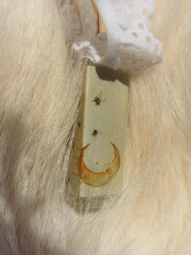 May include: A rectangular, light green glass bottle featuring a printed graphic of a golden crescent moon and several small flying insects. The bottle is topped with a white, decorative lace ribbon tied around the neck of the container.