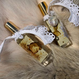 May include: Two rectangular glass perfume bottles with gold caps, each adorned with a white lace ribbon and a small dangling pearl charm. The labels feature a brown illustration of a rabbit and the text HONEYED MOON VIII in black lettering.