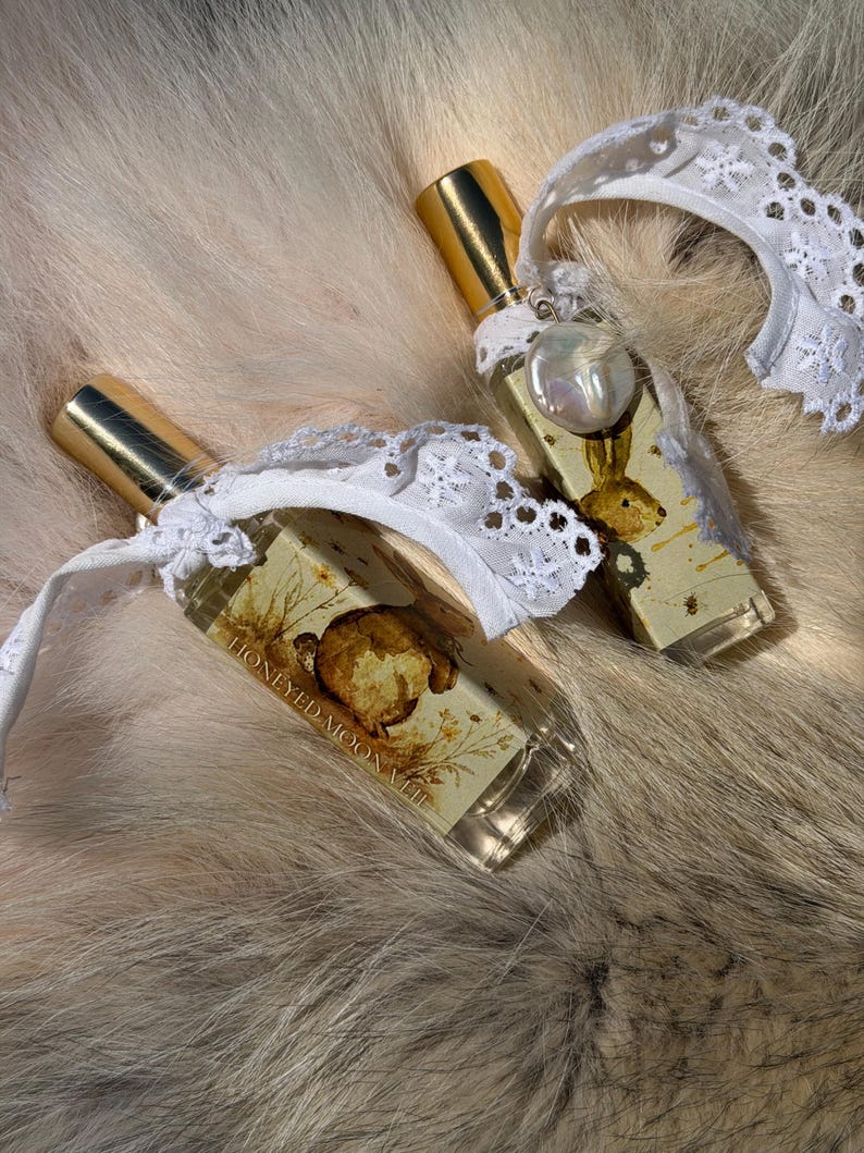 May include: Two rectangular glass perfume bottles with gold caps, each adorned with a white lace ribbon and a small dangling pearl charm. The labels feature a brown illustration of a rabbit and the text HONEYED MOON VIII in black lettering.