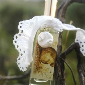May include: A clear glass perfume bottle with a gold cap, featuring a label that reads "EYED MOON VEIL" with a botanical illustration. A white lace ribbon is tied around the neck, adorned with a small, round, white, textured charm.