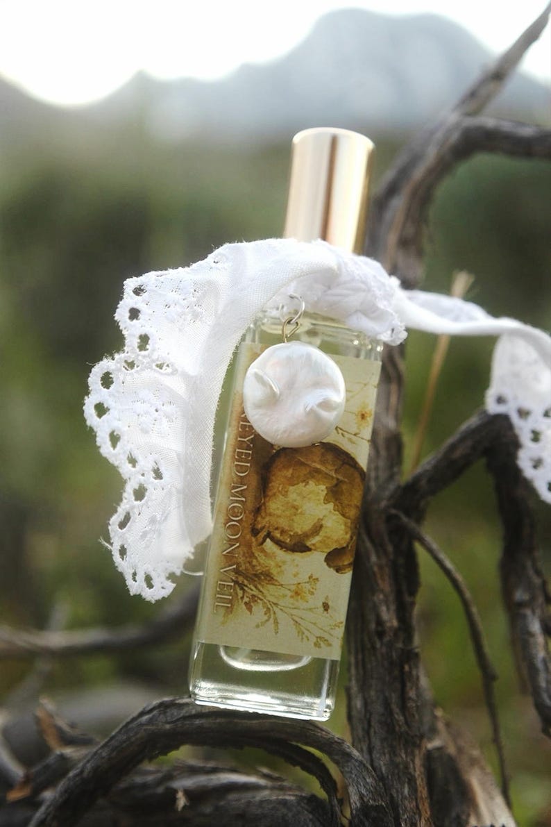 May include: A clear glass perfume bottle with a gold cap, featuring a label that reads "EYED MOON VEIL" with a botanical illustration. A white lace ribbon is tied around the neck, adorned with a small, round, white, textured charm.