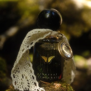 Op de afbeelding: Een donkerglazen parfumflesje met een zwarte bolvormige dop, voorzien van een gouden vlindergrafiek en de tekst Hippies & Cowboys en ethereal melancholy. Een crèmekleurig kanten lint en een kleine ronde metalen hanger zitten om de hals.