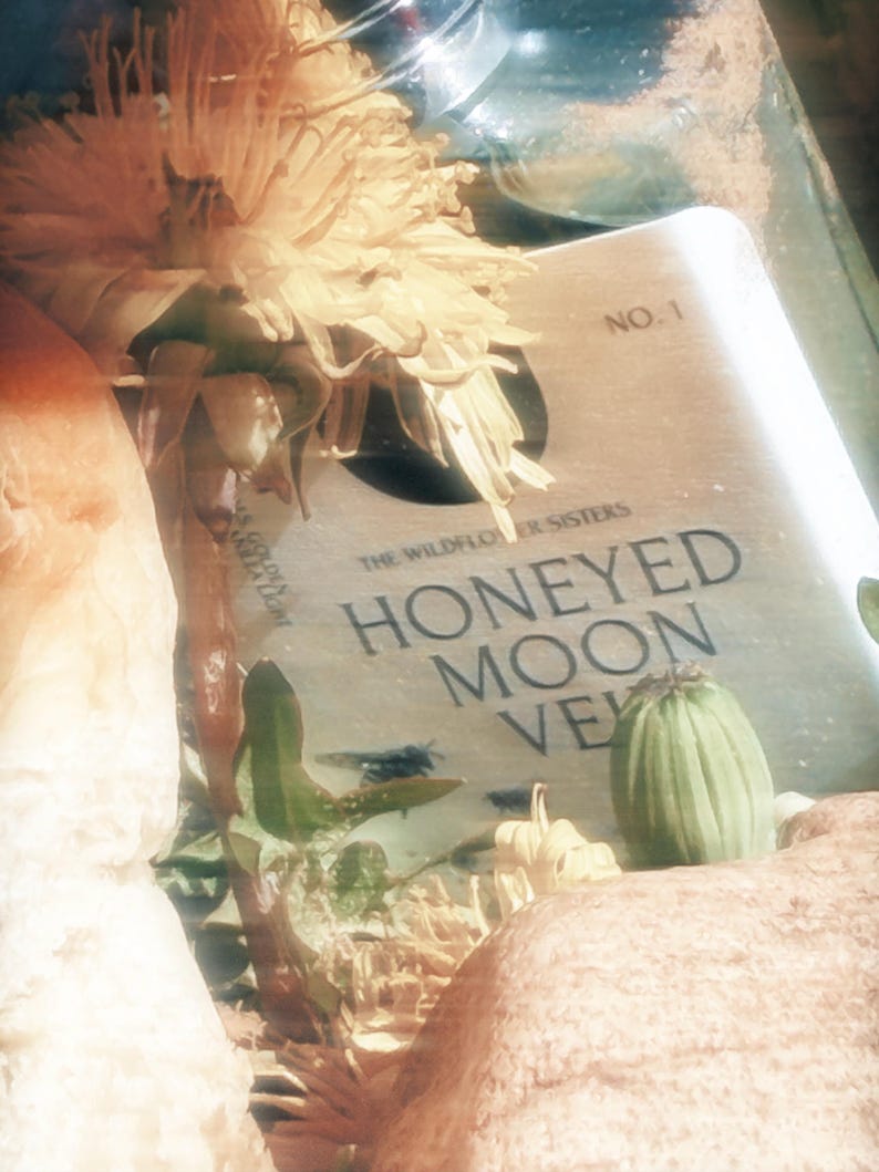 May include: A rectangular label with the text NO. 1, THE WILDFLOWER SISTERS, and HONEYED MOON VEIL, partially obscured by a large yellow dandelion flower, a green seed pod, and textured beige organic elements in a soft, light-filled composition.