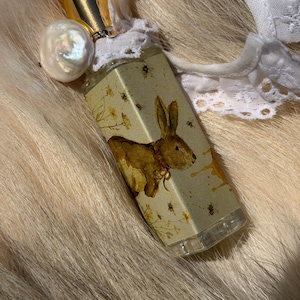 May include: A rectangular glass perfume bottle featuring a label with a brown rabbit illustration, yellow flowers, and small bees. The bottle has a gold cap, a white lace trim around the neck, and a large, iridescent white baroque pearl charm.