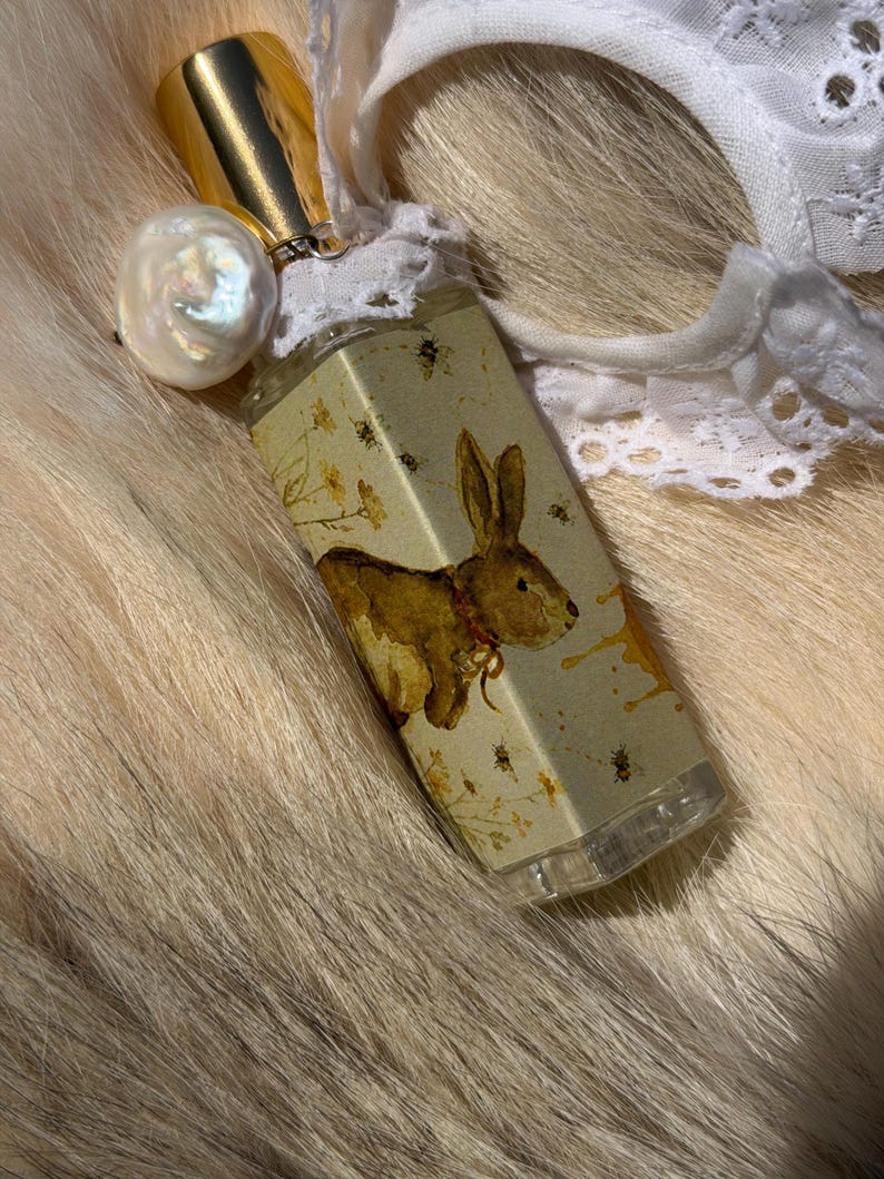 May include: A rectangular glass perfume bottle featuring a label with a brown rabbit illustration, yellow flowers, and small bees. The bottle has a gold cap, a white lace trim around the neck, and a large, iridescent white baroque pearl charm.