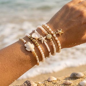 Könnte beinhalten: Ein Set aus fünf geschichteten Armbändern mit weißen Perlen, goldfarbenen Metallakzenten und Anhängern in Form einer Meeresschildkröte, einer Muschel und zwei Seesternen. Die Armbänder sind gestapelt und zeigen ein Küstenthema mit Texturen.