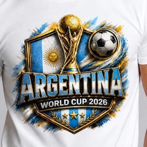 Puede incluir: Una camiseta blanca con un diseño gráfico que incluye el texto ARGENTINA WORLD CUP 2026. El diseño presenta un trofeo dorado, un balón de fútbol, la bandera argentina con su emblema solar y tres estrellas doradas dentro de un escudo decorativo.