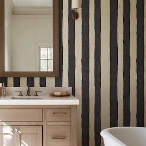Può includere: Un mobile da bagno in legno chiaro con piano bianco e uno specchio rettangolare con cornice marrone. La parete presenta una carta da parati a righe verticali nere e strutturate su fondo crema, con una vasca da bagno bianca visibile.