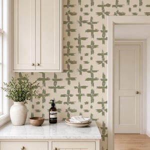 Peut inclure: Papier peint avec un motif répétitif de croix réalisées au pinceau vert olive sur un fond crème. Le design est appliqué sur un mur dans une cuisine avec des armoires couleur crème, un comptoir en marbre et une porte menant à une autre pièce.
