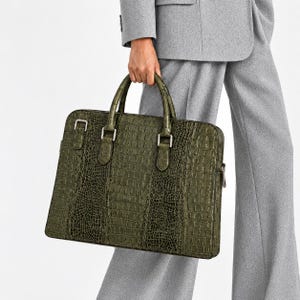 Op de afbeelding: Een rechthoekige olijfgroene aktetas met een gestructureerd krokodillenleerpatroon. De tas is voorzien van twee handvatten aan de bovenzijde, zilverkleurige metalen bevestigingen voor de handvatten en een ritssluiting langs de bovenrand.