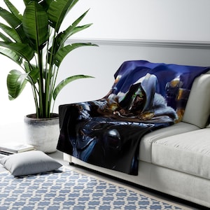 May include: A plush throw blanket featuring a detailed illustration of a character in a hooded cloak with a metallic mask. The artwork is set against a dark, atmospheric background with shades of blue and gold. The blanket is draped over a white sofa.