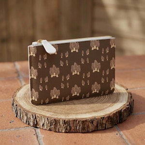 May include: A rectangular brown zippered pouch featuring a repeating pattern of light tan morel mushrooms. The pouch has a white zipper tape and a white pull tab, with a smooth, structured exterior surface and a gold-toned metal zipper slider.