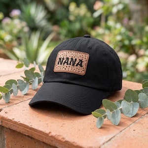 May include: A black baseball cap featuring a rectangular patch on the front. The patch has a tan leopard print background with the word NANA printed in bold black letters across the center. The cap has a curved brim and a structured crown.