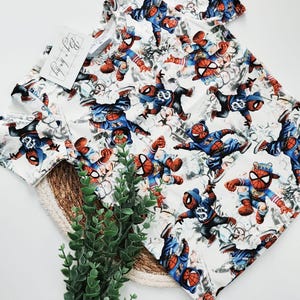 Könnte beinhalten: Weißes Kurzarm-T-Shirt mit einem sich wiederholenden Muster eines Cartoon-Spider-Man-Charakters in verschiedenen Action-Posen. Die Figur trägt ein blaues und rotes Kostüm mit einer Spider-Man-Maske. Das T-Shirt hat ein Etikett mit der Aufschrift "By Baby".
