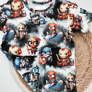 Könnte beinhalten: Ein kurzärmeliges T-Shirt mit einem sich wiederholenden Muster von Cartoon-Superhelden, darunter Iron Man, Captain America, Batman und Spider-Man, vor einem schwarz-weißen Wolkenhintergrund. Das Shirt hat ein Etikett mit der Aufschrift "By baby".