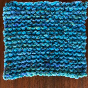 May include: A blue knitted square with a textured surface. The yarn is a mix of blue and green shades with some brown flecks.
