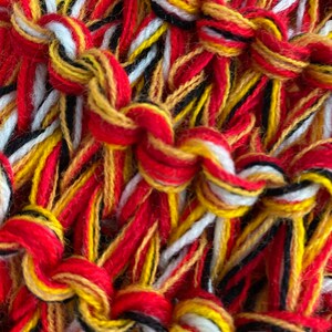 May include: Close-up of a woven textile with red, white, yellow, and black threads. The threads are woven together in a complex pattern, creating a textured surface.