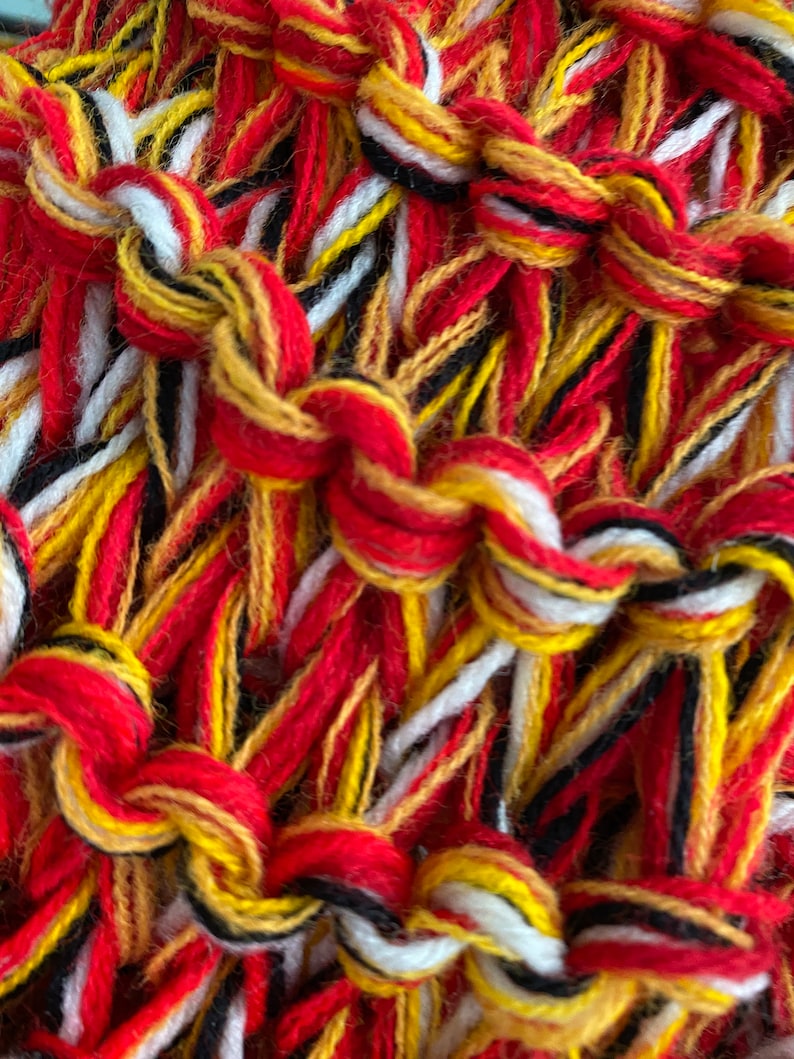 May include: Close-up of a woven textile with red, white, yellow, and black threads. The threads are woven together in a complex pattern, creating a textured surface.