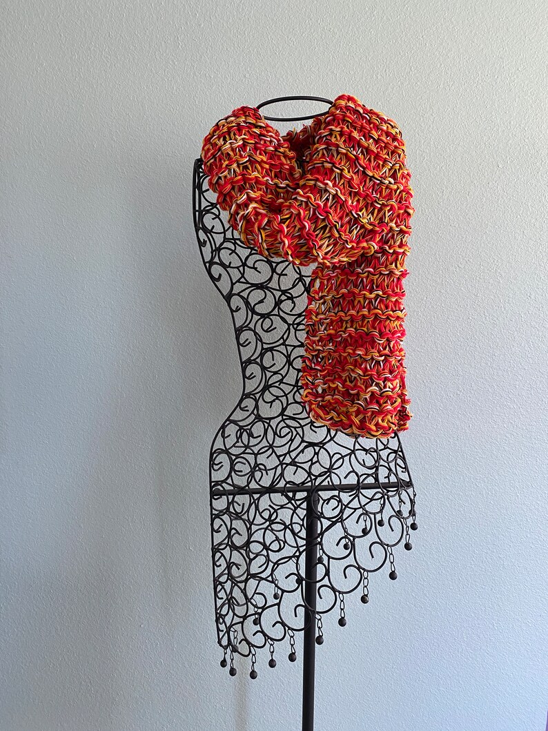 May include: A red, yellow and white knitted scarf displayed on a black metal mannequin.