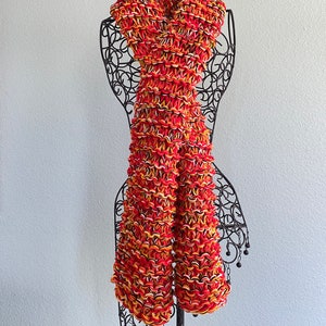 May include: A red, orange and white knitted scarf with a textured pattern. The scarf is displayed on a black metal mannequin.