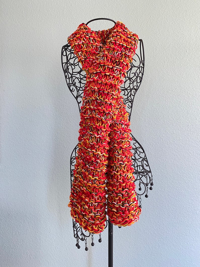 May include: A red, orange and white knitted scarf with a textured pattern. The scarf is displayed on a black metal mannequin.