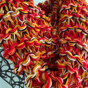 May include: A close-up of a knitted scarf in red, white, black, and yellow yarn. The scarf is made with a chunky yarn and has a textured, woven look.