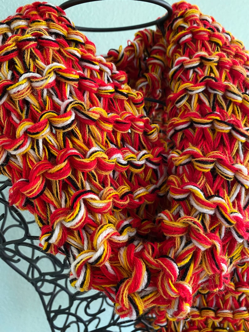 May include: A close-up of a knitted scarf in red, white, black, and yellow yarn. The scarf is made with a chunky yarn and has a textured, woven look.