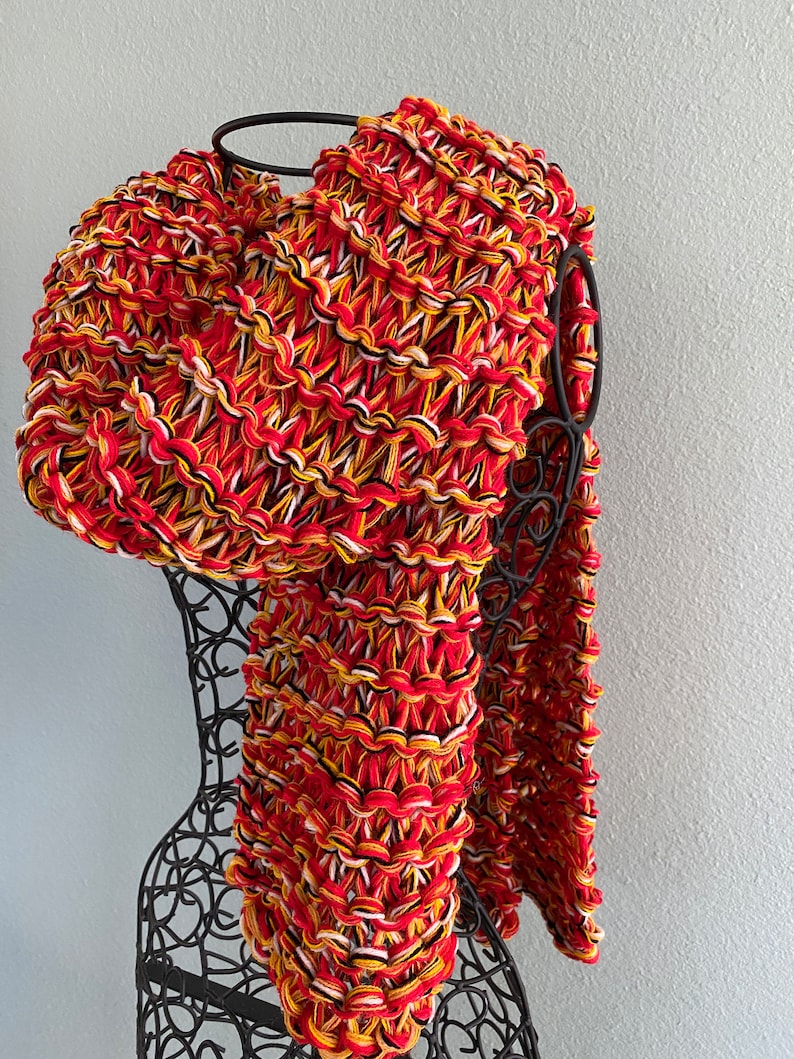 May include: A chunky knit scarf in red, yellow, black and white. The scarf is draped over a black metal mannequin.