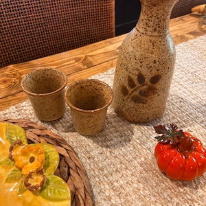 May include: A ceramic sake set featuring a tall, speckled beige carafe with a cork stopper and two matching small cups, each decorated with a brown leaf motif. Beside the set sits a small, glossy orange pumpkin figurine with a dark leaf.