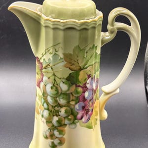 May include: A tall, fluted ceramic coffee pot featuring a light green and yellow gradient base. The body is decorated with a painted cluster of green and purple grapes with green leaves. It includes a matching lid with a finial and a curved handle.