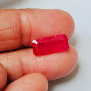 May include: A rectangular, faceted gemstone with a vibrant, saturated red color. The stone features a clear, polished surface with sharp edges and a classic emerald-style cut, displaying internal light reflections and a deep, uniform crimson hue.