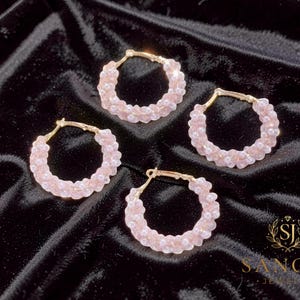 Puede incluir: Cuatro pendientes de aro con estructura de metal dorado adornados con pequeñas cuentas translúcidas de color rosa claro y diminutas perlas blancas dispuestas en un patrón trenzado. Las piezas son circulares y tienen cierre de bisagra.