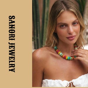 Puede incluir: Un collar que presenta una serie de cuentas lisas, redondas y multicolores en tonos rosa, amarillo, turquesa y verde, intercaladas con una cuenta de tono dorado. El texto SANORI JEWELRY aparece verticalmente a la izquierda.