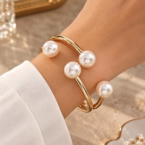 Puede incluir: Una pulsera de brazalete abierto en tono dorado con una banda de metal lisa y pulida que se envuelve alrededor de la muñeca. Los extremos de la pulsera están adornados con tres perlas artificiales blancas redondas de distintos tamaños.
