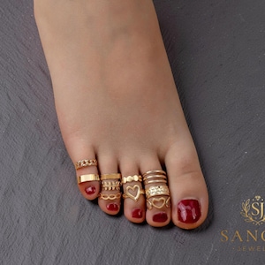 Puede incluir: Una colección de anillos para los dedos de los pies en tono dorado, con varios diseños que incluyen un patrón de cadena, una banda lisa, una forma de corazón y anillos apilados. Las uñas están pintadas de rojo oscuro y el logo de Sanori Jewelry es visible.