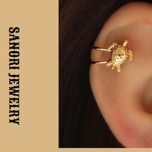 May include: A gold-toned ear cuff featuring a small turtle design with a textured shell. The jewelry piece is positioned on the cartilage of an ear. The text SANORI JEWELRY is printed vertically in black on the left side of the image.