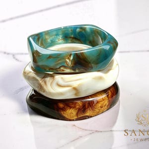 May include: A stack of three chunky resin bangles with irregular, rounded square shapes. The top bangle is teal and brown, the middle is cream and beige, and the bottom is dark amber and brown. Each piece features a swirling, marbled pattern.