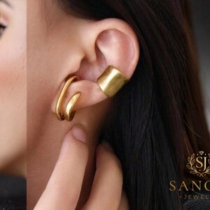 Puede incluir: Un par de pendientes tipo ear cuff dorados. Uno es una banda ancha, plana y lisa para el cartílago superior, mientras que el otro es un diseño de alambre grueso, redondeado y abierto para el lóbulo. La joyería tiene un acabado pulido.