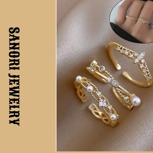 Könnte beinhalten: Drei goldfarbene offene Ringe mit filigranen Bändern, kleinen weißen Perlen und klaren funkelnden Steinen. Ein Ring zeigt eine kreuzförmige Anordnung von Steinen. Der Text SANORI JEWELRY ist auf der linken Seite des Bildes zu sehen.