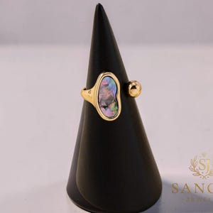 May include: A gold ring featuring an irregular, organic-shaped setting inlaid with a shimmering, iridescent abalone shell inlay. The band is open-style, finished with a small, smooth gold sphere on one side and the shell inlay on the other.