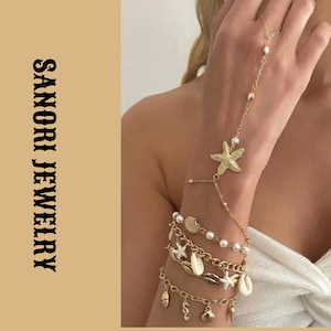 Peut inclure: Un bracelet de main doré avec une breloque étoile de mer centrale, relié par une fine chaîne à une bague. La partie poignet comprend plusieurs chaînes superposées ornées de petites perles blanches, de divers coquillages et d'une petite breloque hippocampe. Le texte indique SANORI JEWELRY.
