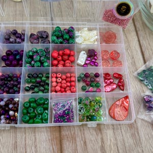 May include: A clear plastic storage container with twenty-four compartments filled with an assortment of colorful beads in various shapes, sizes, and textures, including purple, green, red, and white varieties, alongside several small plastic bags of loose beads.