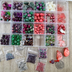 May include: A clear plastic organizer box filled with various colorful beads in shades of green, pink, purple, red, and white. Below the box are several small plastic bags containing assorted seed beads and two large red shell-like pieces.