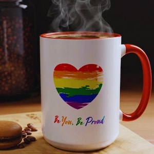 May include: A white ceramic mug with a red handle and rim, featuring a rainbow heart design. The mug has the text "Be You, Be Proud" in colorful letters. Steam rises from the coffee inside the mug.