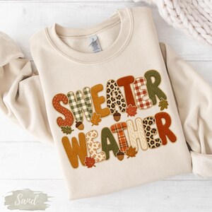 May include: A sand-colored crewneck sweatshirt featuring the text "SWEATER WEATHER" in large, multi-patterned letters. The design incorporates autumn-themed motifs including leopard print, plaid, floral patterns, and small brown acorn and leaf icons.