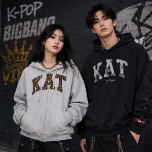 Puede incluir: Una sudadera gris claro con cremallera y logo KAT de estampado de leopardo y una sudadera negra con capucha y logo KAT by Nagan de camuflaje gris, con detalles de cordones en las mangas y un pequeño parche rectangular rojo y blanco en el puño.